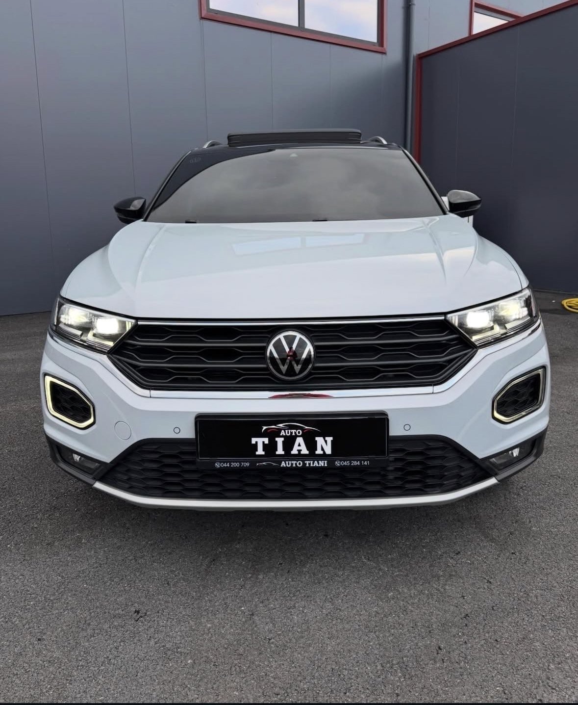 White Volkswagen T-Roc SUV parked in front of gray building showing front view with LED headlights and black trim