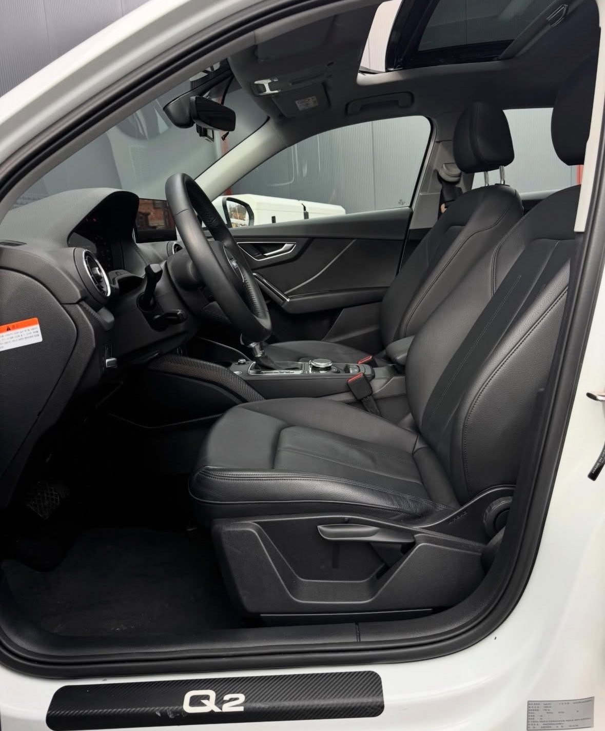 Interior of white Audi Q2 with black leather seats, steering wheel, and dashboard viewed from drivers side door open