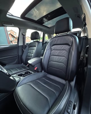 Interior view of a vehicle showing black leather front seats with white stitching, panoramic sunroof, and dashboard with cloudy sky visible outside windows