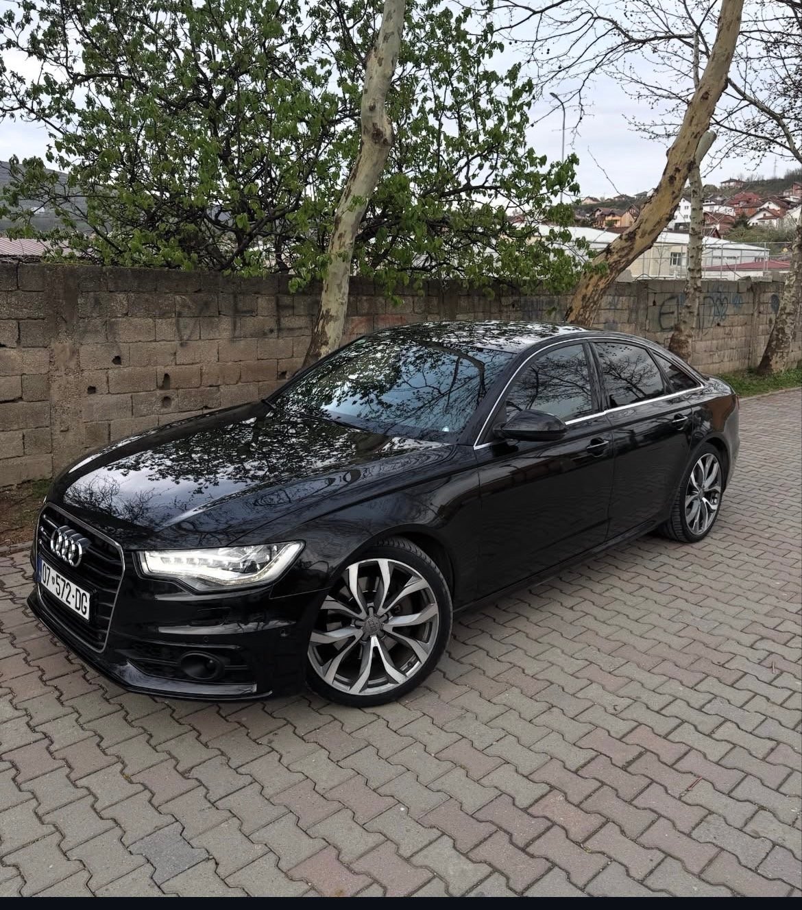 Black Audi sedan parked on brick driveway against stone wall with trees in background
