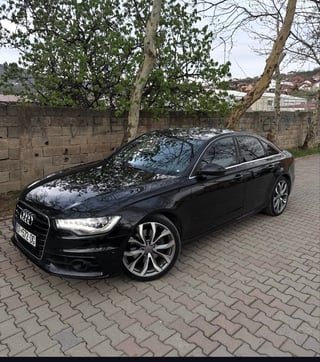 Black Audi sedan parked on brick driveway against stone wall with trees in background